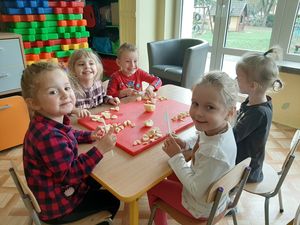 Zdjęcie przedstawia dzieci siedzące na krzesłach przy stoliku w przedszkolnej sali. W rekach trzymają plastikowe noże, przed nimi leży duża deska do krojenia, a na niej pokrojone na małe części owoce – banany i jabłka.