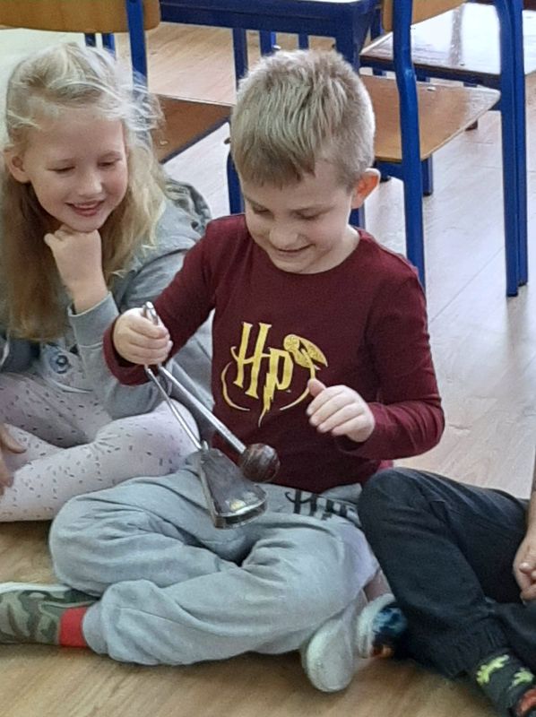 Zdjęcie przedstawia dwójkę przedszkolaków – chłopca i dziewczynkę z grupy Skowronki. Dzieci siedzą na podłodze w przedszkolnej sali, chłopiec trzyma w rękach instrument perkusyjny o nazwie vibraslap.