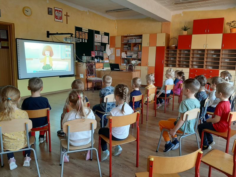 Zdjęcie przedstawia czterolatki, które siedzą w przedszkolnej sali na krzesłach. Dzieci oglądają film wyświetlany na ekranie.