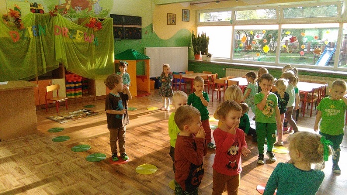 Zdjęcie przedstawia trzyletnie dzieci, które stoją w rozsypce w przedszkolnej sali. Na swoich ustach trzymają palce w geście ciszy.