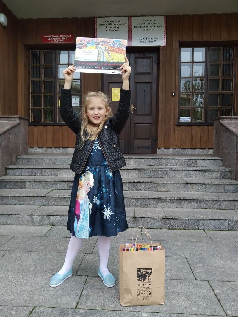 Zdjęcie przedstawia sześcioletnią dziewczynkę trzymającą w rękach dyplom za udział w konkursie. W tle schody i drzwi wejściowe do Muzeum Kultury Białoruskiej w Hajnówce.