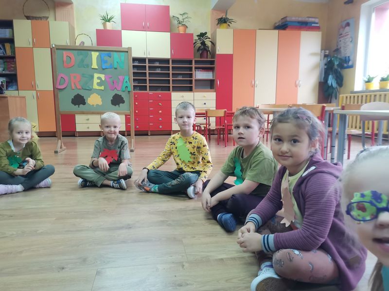 Fotografia przedstawia dzieci siedzące w kole na podłodze w przedszkolnej sali. Na bluzkach mają emblematy liści klonu i dębu w różnych kolorach.