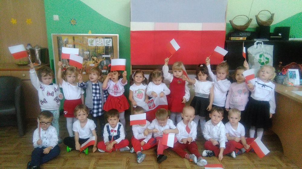 Na zdjęciu dzieci trzyletnie pozują do zdjęcia. Każde trzyma w ręku papierową biało-czerwoną flagę. Z tyłu na ścianie, duża flaga Polski.