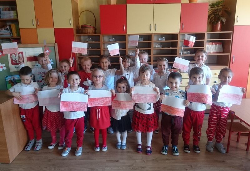 Zdjęcie przedstawia dzieci stojące w dwóch rzędach w przedszkolnej sali, w rękach trzymają rysunki flagi Polski.