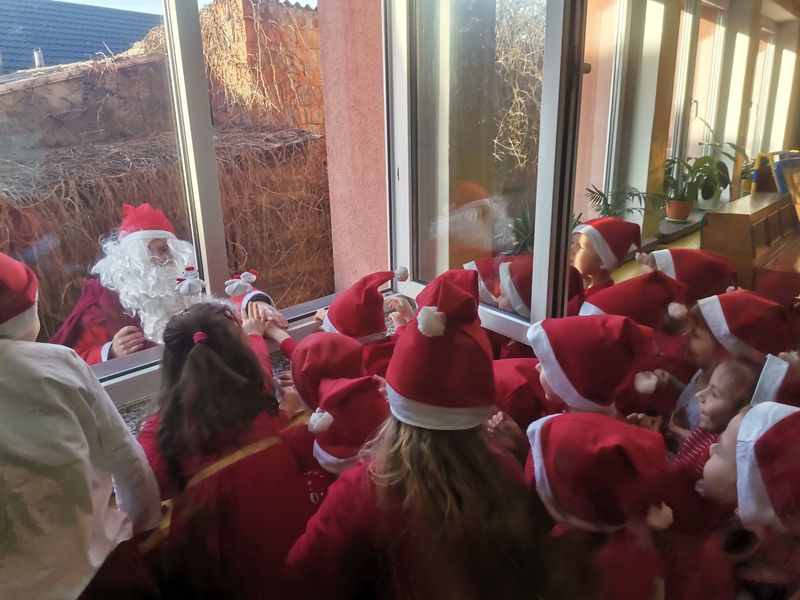 Zdjęcie przedstawia gromadkę dzieci ubranych na czerwono w czapkach Mikołaja. Za oknem jest święty Mikołaj . Dzieci przez okno machają do niego,a on przybija im piątkę.
