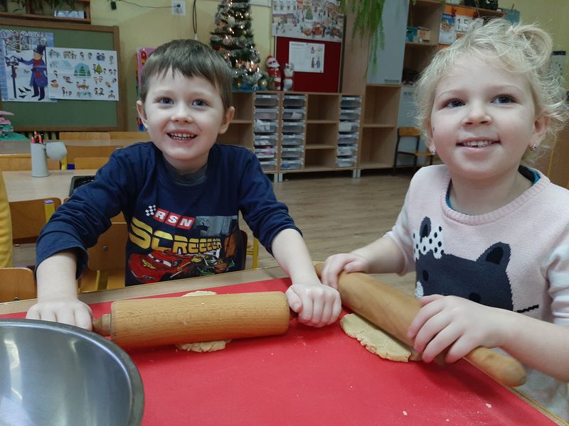 Na zdjęciu dwoje dzieci siedzi przy stole w przedszkolnej sali. W rękach trzymają kuchenne wałki, wałkują ciasto na pierniczki.