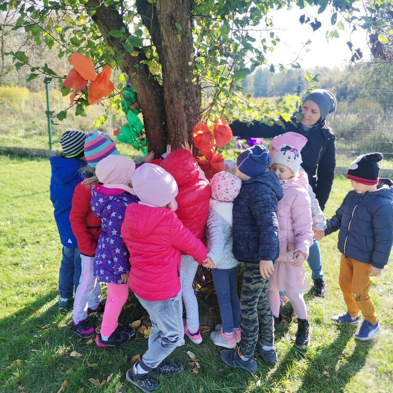 Dzieci stoją wkoło drzewa i dotykają je przekazując życzenia z okazji Dnia Drzewa. 
