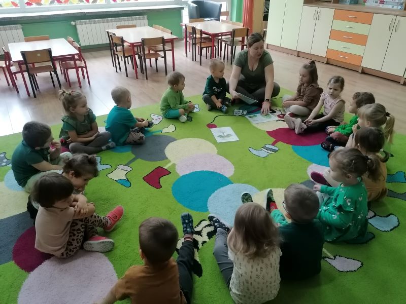 Dzieci w sali przedszkolnej siedzą na dywanie w koło