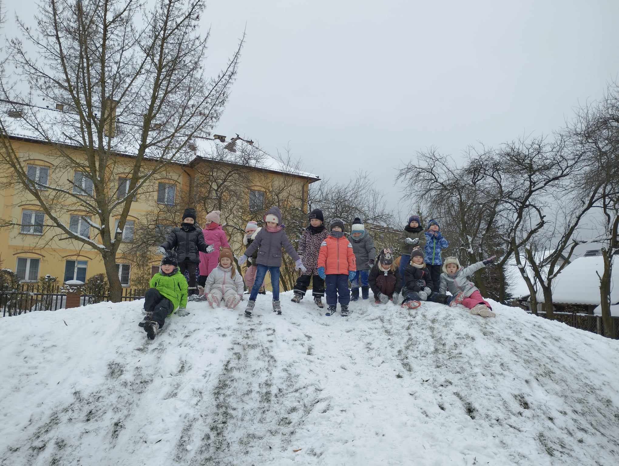 Na zdjęciu dzieci znajdują się na zaśnieżonej górce w ogrodzie przedszkolnym. 