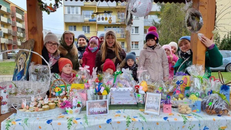 Dzieci,rodzice i nauczycielki stoją przy stoisku Kolebki „podczas” Kiermaszu Wielkanocnego. Na stole znajdują się ręcznie wykonane ozdoby i wypieki wielkanocne. 