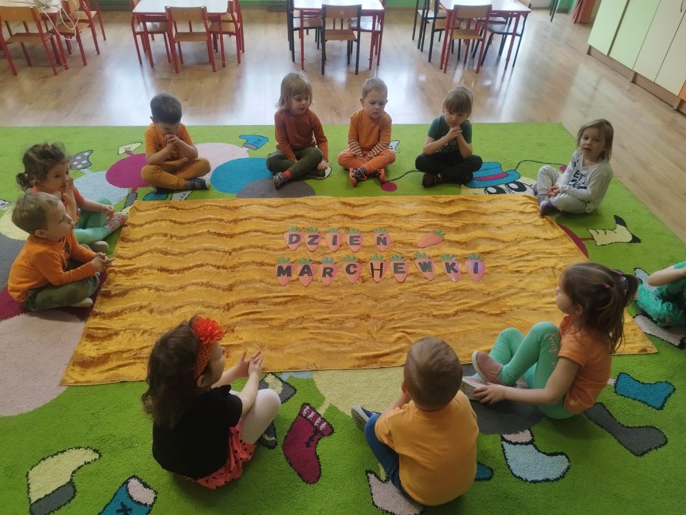 Dzieci siedzą na dywanie wokół pomarańczowego koca. Na środku napis: „Dzień Marchewki”.