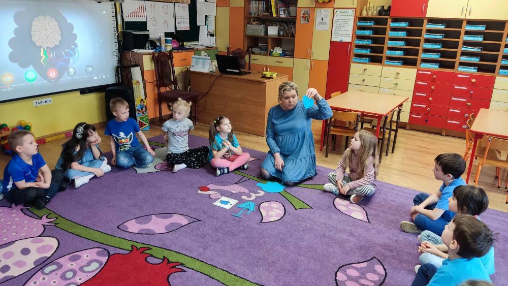 Grupa przedszkolaków wraz z panią pedagog siedzą na dywanie podczas zajęć.